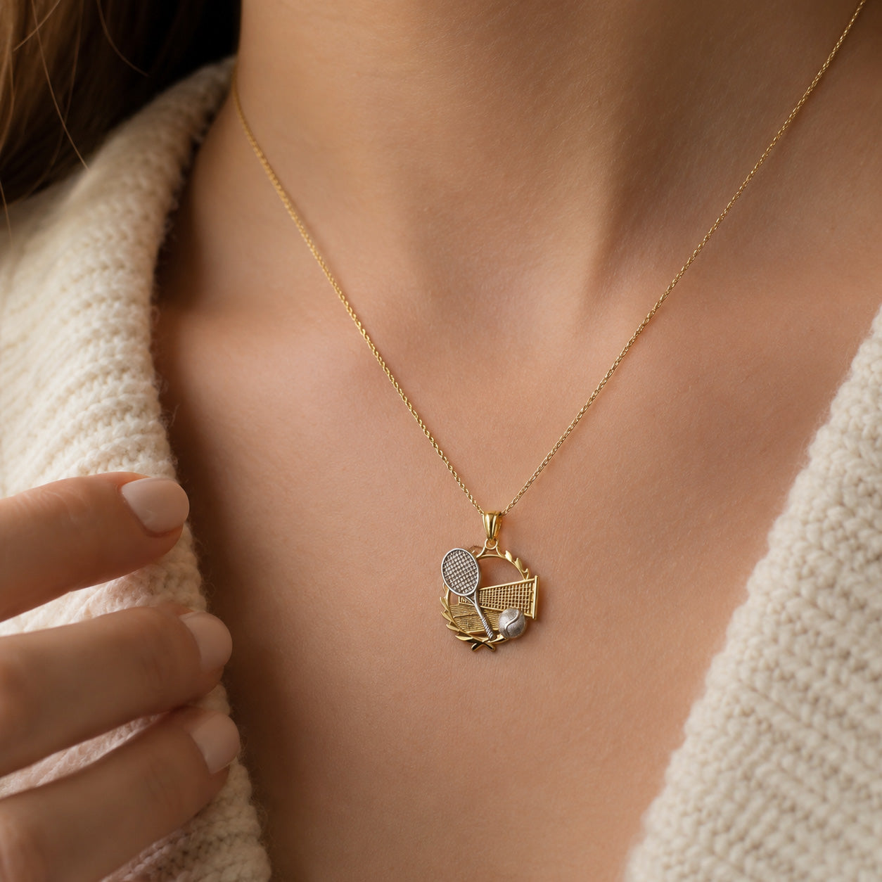 [group: 10k-gold]Gold necklace with a tennis-themed pendant on a person wearing a beige sweater.