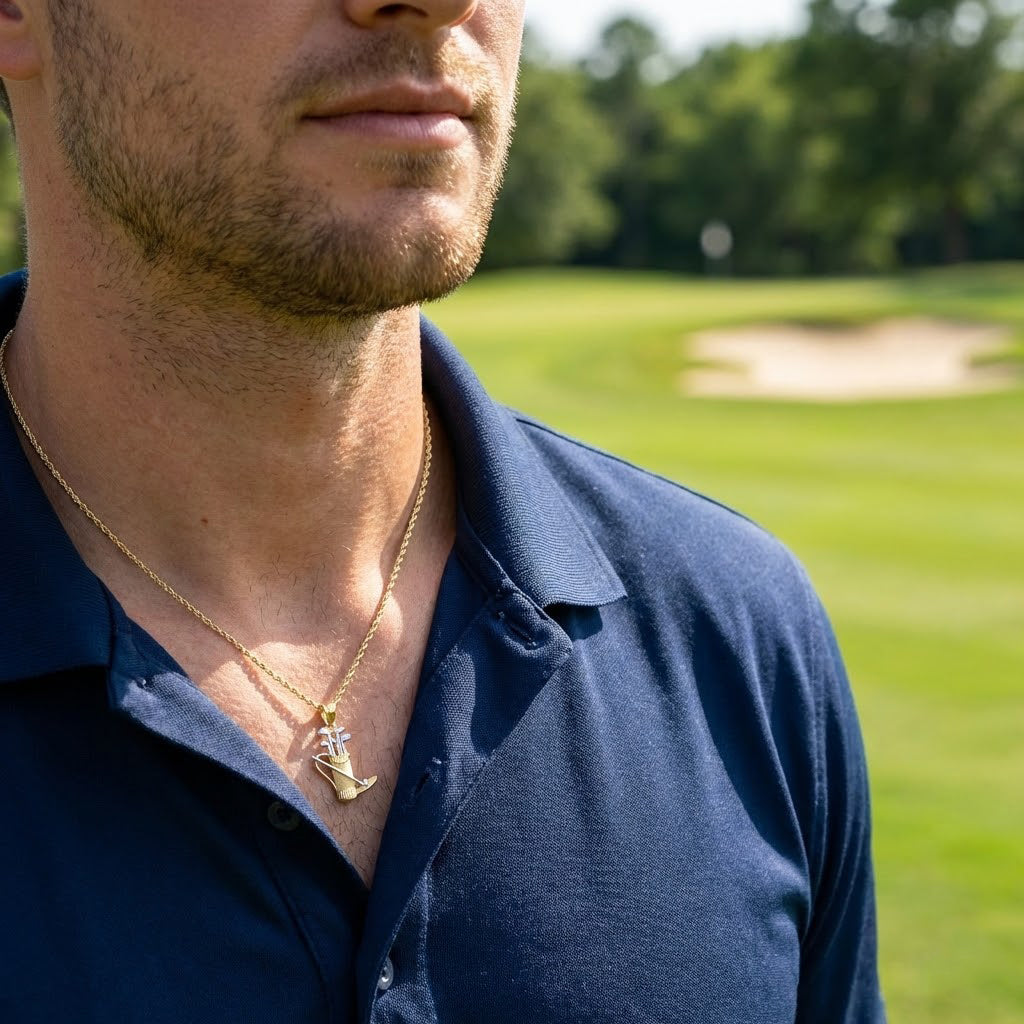 A close-up lifestyle shot of a man wearing a gold chain necklace with a small gold golf ball pendant, wearing a navy blue polo shirt on a golf course.