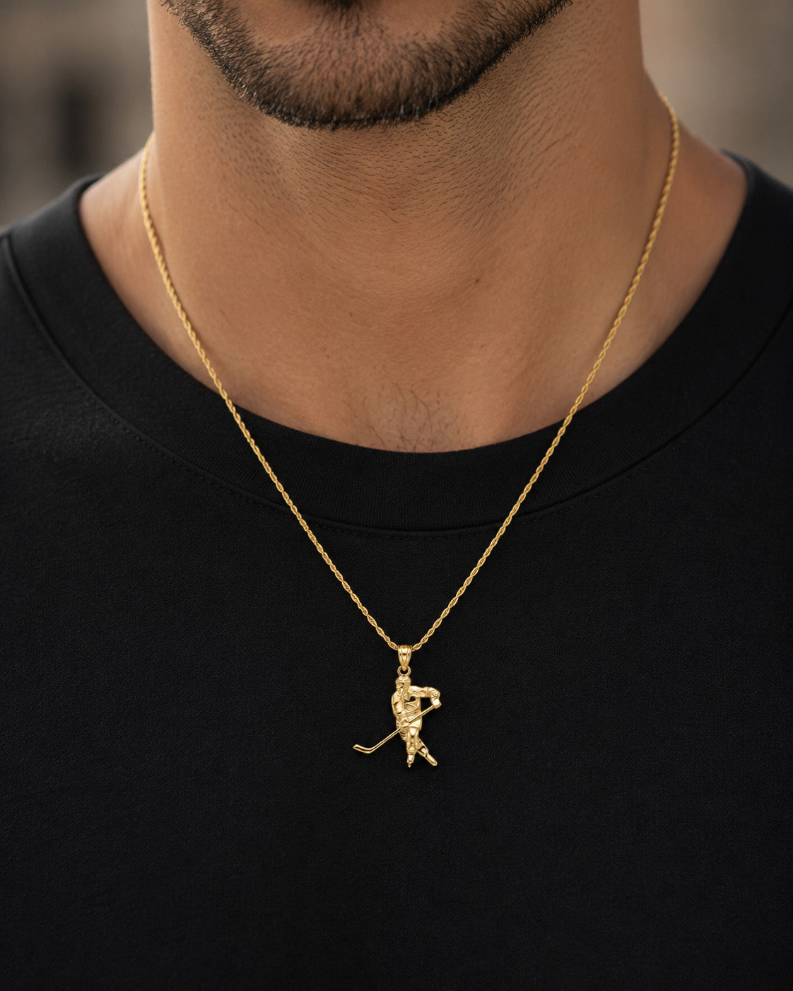 A close-up of a man's neck and chest wearing a gold chain necklace with a golfer pendant, paired with a black t-shirt.