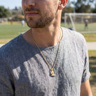 Man wearing a gold necklace with a soccer pendant outdoors on a grassy field.