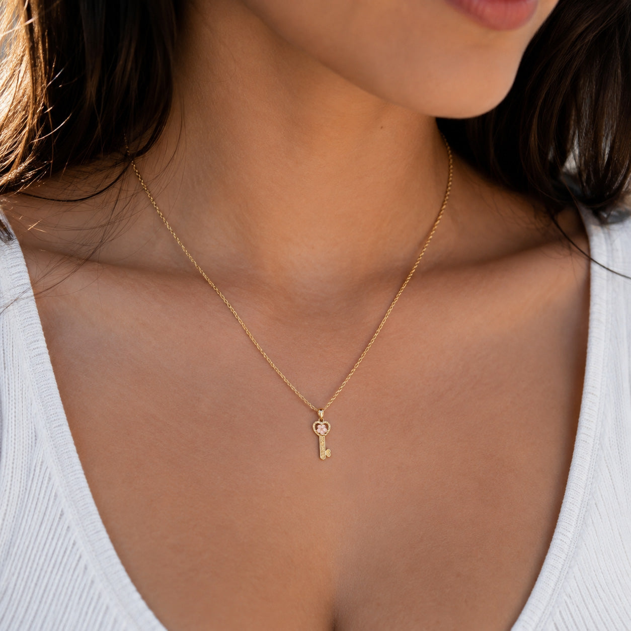 A close-up lifestyle shot of a model wearing a delicate gold chain necklace with a small key-shaped pendant, styled with a white ribbed top.