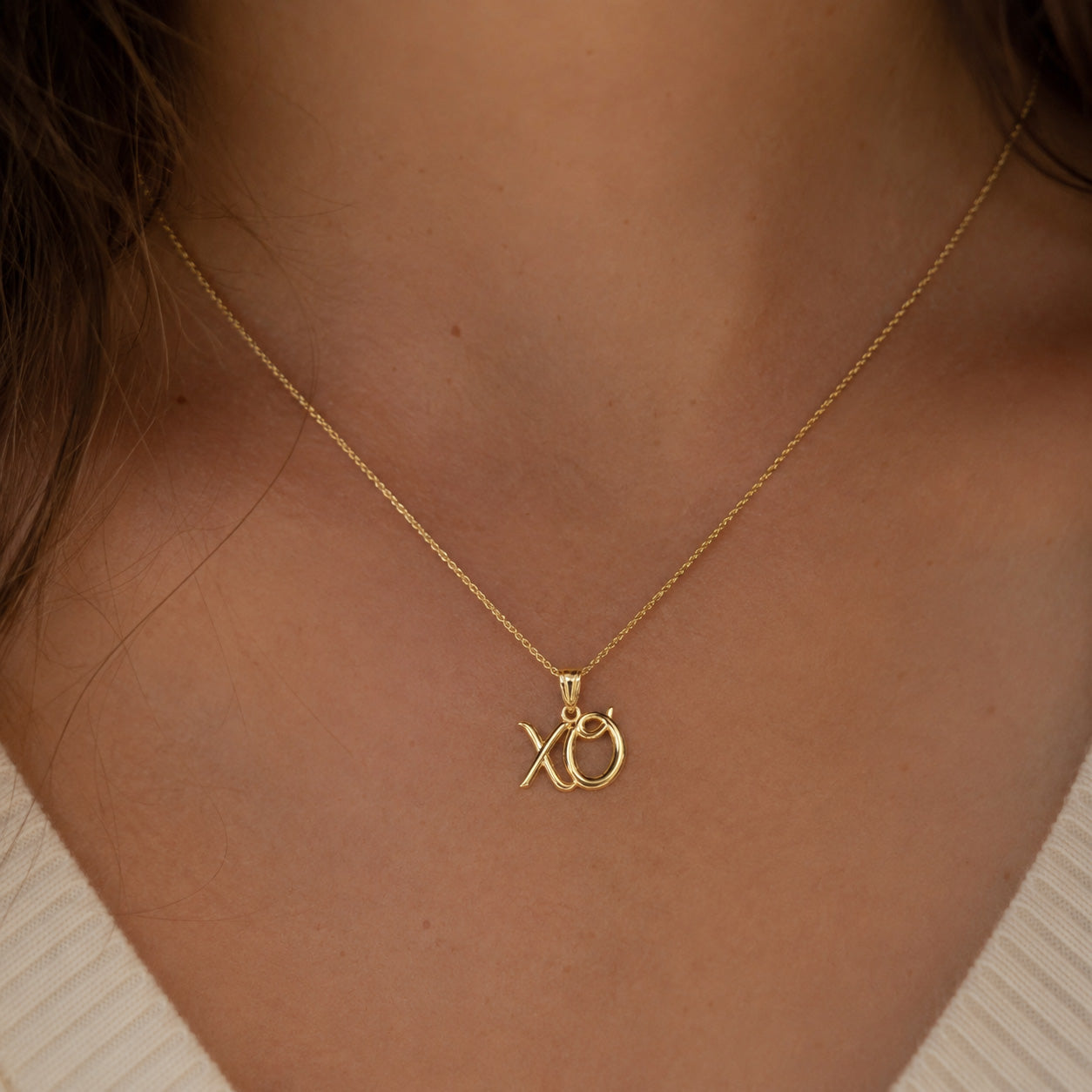 A close-up of a person's neck and chest wearing a delicate gold chain necklace with an interlocking 'XO' pendant.