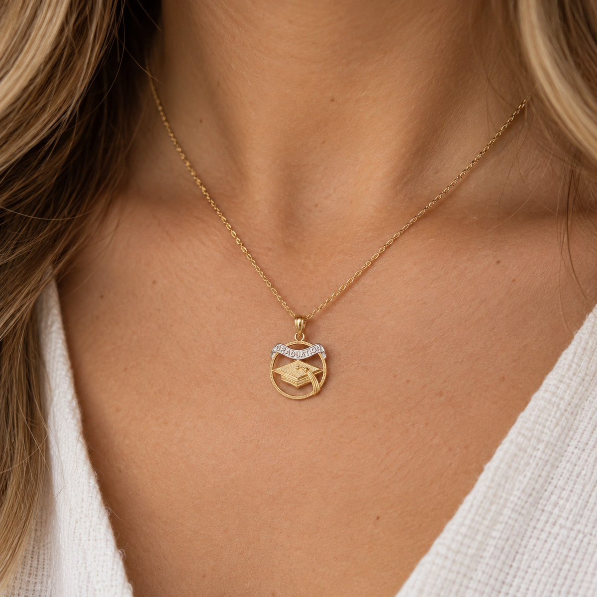 A close-up of a woman wearing a delicate gold chain necklace with a circular pendant featuring a graduation cap design and a small silver accent at the top.