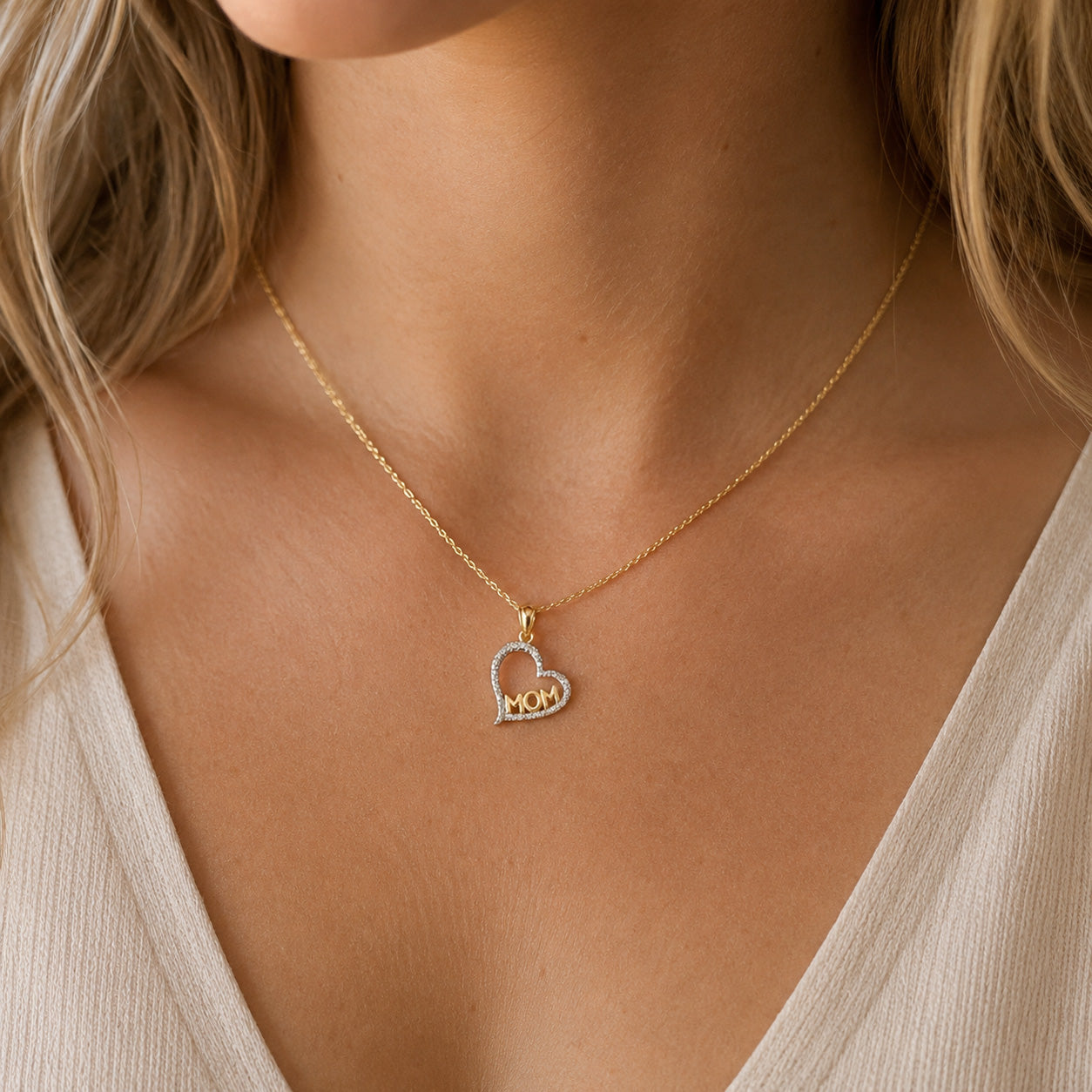 A close-up of a woman's neck and chest wearing a delicate gold chain necklace with a heart-shaped pendant. The pendant features the word 'MOM' in gold letters, with the left side outlined in small clear stones.